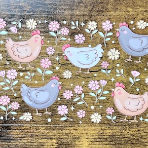 Puede incluir: Una hoja de pegatinas decorativas con pollos de dibujos animados en varios colores, incluyendo azul y melocotón, intercalados con flores rosas y blancas y hojas verdes. El fondo es un patrón de vetas de madera.