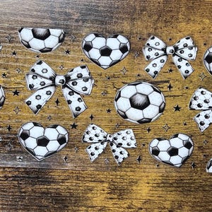 May include: A collection of soccer-themed stickers featuring soccer balls, heart-shaped soccer balls, and bows with soccer ball patterns. The stickers are in black and white, with a wood-grain background.