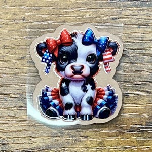 May include: A sticker featuring a cartoon cow with patriotic red, white, and blue accents. The cow has a black and white coat, bows, and a tutu-like skirt. Stars and stripes decorate the bows and skirt.