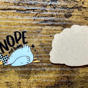 May include: A sticker featuring a light blue duck with a checkered bow, the words "NOPE not today", and a blank, cloud-shaped sticker. The duck is lying down on a wooden surface.
