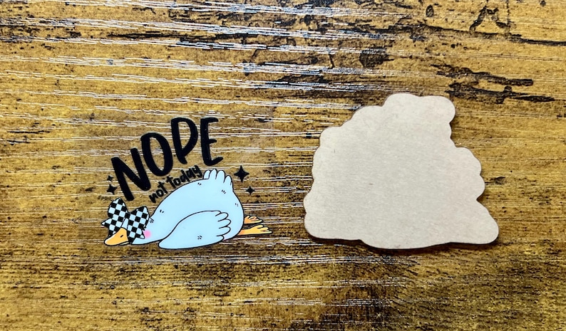 May include: A sticker featuring a light blue duck with a checkered bow, the words "NOPE not today", and a blank, cloud-shaped sticker. The duck is lying down on a wooden surface.