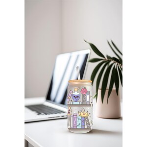 May include: A glass tumbler with a bamboo lid and straw, decorated with a whimsical bookshelf design. The illustration features books, potion bottles, a crown, and a rose in pastel colors. The tumbler is on a white surface with a laptop and plant in the background.