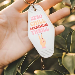 Puede incluir: Llavero blanco de motel con el texto "Zero Chill Maximum Thrill" en letras rosa, rojo y amarillo. Un signo de paz con uñas rosas está debajo del texto.