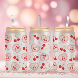 May include: Three frosted glass tumblers with cherry and heart patterns. Each tumbler has a bamboo lid and a silver straw. The background is a soft pink with bokeh lights, creating a sweet, romantic vibe.