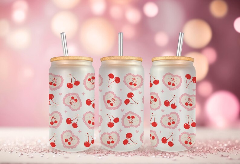 May include: Three frosted glass tumblers with cherry and heart patterns. Each tumbler has a bamboo lid and a silver straw. The background is a soft pink with bokeh lights, creating a sweet, romantic vibe.