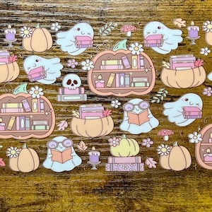 Könnte beinhalten: Eine Sammlung skurriler Halloween-Sticker. Die Sticker zeigen Geister beim Lesen von Büchern, Kürbisse, Bücherregale, Totenköpfe, Kerzen und florale Akzente. Die Farbpalette umfasst Pastelltöne von Orange, Rosa und Blau.