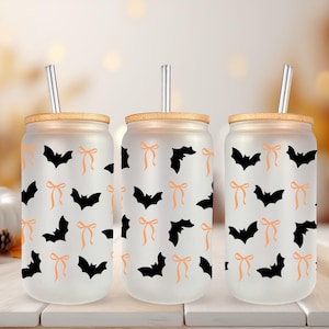 May include: Three frosted glass tumblers with bamboo lids and silver straws. Each tumbler features a repeating pattern of black bats and orange bows. The tumblers are designed for cold beverages and are perfect for Halloween.
