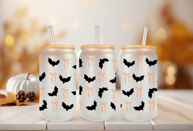 May include: Three frosted glass tumblers with bamboo lids and silver straws. Each tumbler features a repeating pattern of black bats and orange bows. The tumblers are designed for cold beverages and are perfect for Halloween.