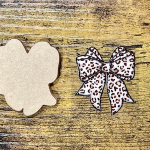 May include: Two wooden craft shapes on a wood grain surface. One is a blank, light brown shape. The other is a bow with a white background and a brown leopard print pattern. The bow has a black outline.