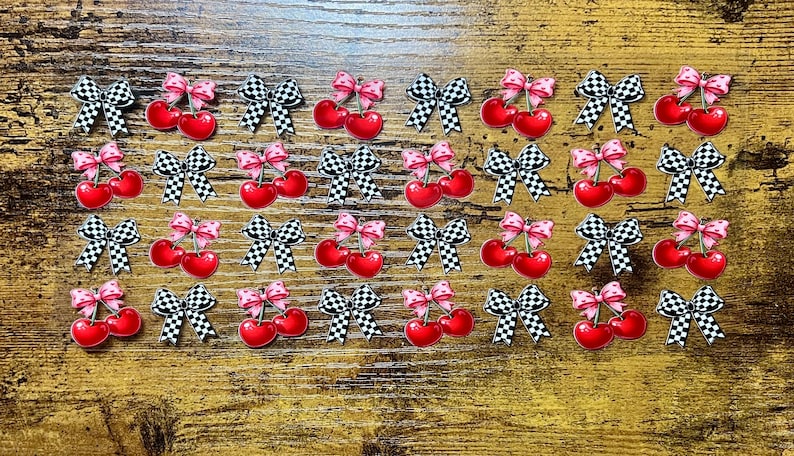 Pu&ograve; includere: Una collezione di ciondoli decorativi piatti disposti in una griglia. Ogni ciondolo presenta una coppia di ciliegie rosso brillante sormontate da un fiocco a pois rosa e bianchi o una coppia di ciliegie con un fiocco a scacchi bianchi e neri.
