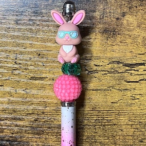 May include: A decorative pen with a pink and white speckled barrel. The pen is topped with a pink bunny wearing blue glasses, a green bead, and a pink textured ball.