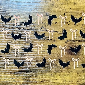 May include: A collection of black bat-shaped cutouts and light pink bow-shaped cutouts arranged on a wooden surface. The bats and bows are evenly spaced, creating a decorative pattern. The bats are solid black, and the bows are a pale pink.