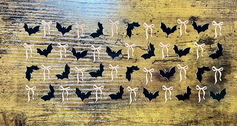 May include: A collection of black bat-shaped cutouts and light pink bow-shaped cutouts arranged on a wooden surface. The bats and bows are evenly spaced, creating a decorative pattern. The bats are solid black, and the bows are a pale pink.