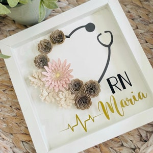 Puede incluir: Una caja de sombra enmarcada en blanco con un diseño de estetoscopio negro y flores de papel en tonos marrones y rosas. El texto "RN Maria" y un gráfico de latido del corazón dorado también están incluidos.
