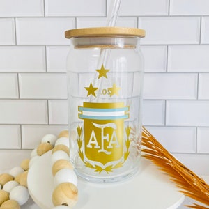 May include: A clear glass jar with a bamboo lid and a straw. The jar has a gold design of a crest with three stars and the letters "AFA" inside. The jar is sitting on a white pedestal.
