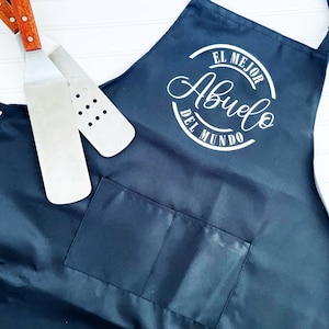K&ouml;nnte beinhalten: Schwarze Sch&uuml;rze mit einem wei&szlig;en Kreis und dem Text "El Mejor Abuelo Del Mundo" in kursiv. Die Sch&uuml;rze hat eine gro&szlig;e Tasche auf der Vorderseite.