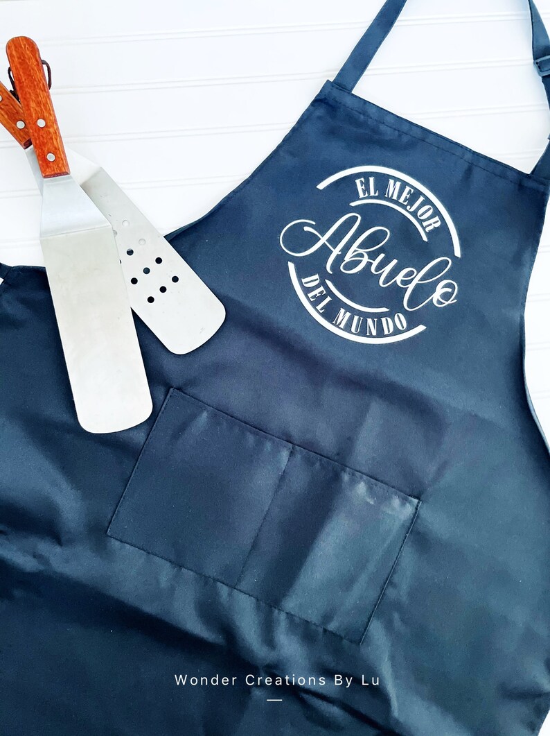 K&ouml;nnte beinhalten: Schwarze Sch&uuml;rze mit einem wei&szlig;en Kreis und dem Text "El Mejor Abuelo Del Mundo" in kursiv. Die Sch&uuml;rze hat eine gro&szlig;e Tasche auf der Vorderseite.
