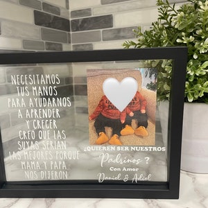 May include: A black picture frame with a white heart cutout and a photo of two children in red and black outfits. The text in the frame reads "Necesitamos tus manos para ayudarnos a aprender y crecer. Creo que las suyas serian las mejores porque mama y papa nos dijeron ¿Quieren ser nuestros Padrinos? Con Amor Daniel & Adiel"