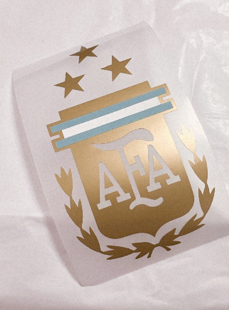 May include: Gold and blue vinyl decal of the Argentine Football Association (AFA) logo. The logo features a shield with the letters "AFA" inside, two horizontal blue stripes, and three gold stars above the shield. The shield is surrounded by a laurel wreath.
