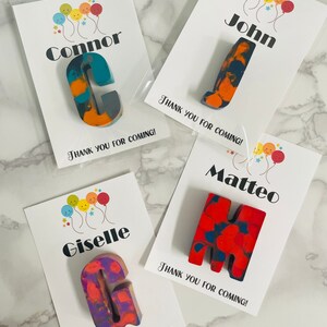 May include: Four personalized party favor bags with colorful crayon letters and thank you messages. The bags feature the names Connor, John, Matteo, and Giselle.