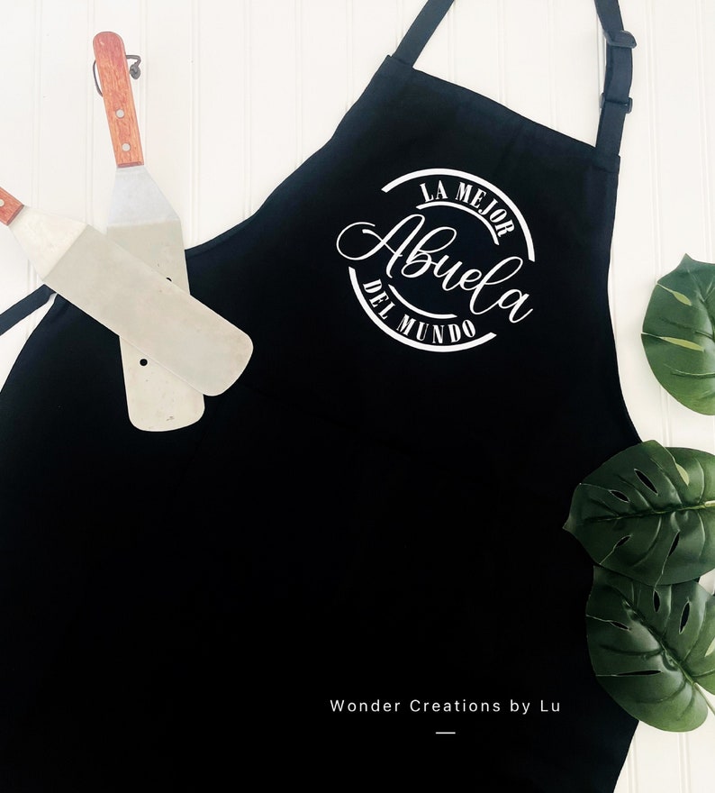 K&ouml;nnte beinhalten: Schwarze Sch&uuml;rze mit einem wei&szlig;en Kreisdesign, auf dem "La Mejor Abuela Del Mundo" auf Spanisch steht. Die Sch&uuml;rze h&auml;ngt vor einem wei&szlig;en Hintergrund.