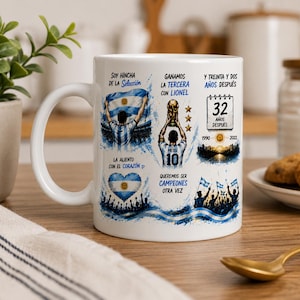 May include: A white ceramic mug featuring blue and white graphics related to the Argentina national soccer team. The design includes illustrations of a player holding a trophy, a heart-shaped flag, and text in Spanish celebrating a 32-year victory.