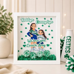 May include: A framed artwork featuring two cartoon cheerleaders in green and blue uniforms, with the text "We love you Coach Amy." The artwork includes green shamrocks and pom-poms, and the names "Katelyn" and "Evangelyn" are displayed at the top.