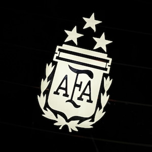 以下が含まれることがあります： 盾の中に「AFA」の文字が入った銀色のエンブレム。3つの星が上にあり、月桂樹の冠で囲まれています。エンブレムは黒い背景に配置されています。