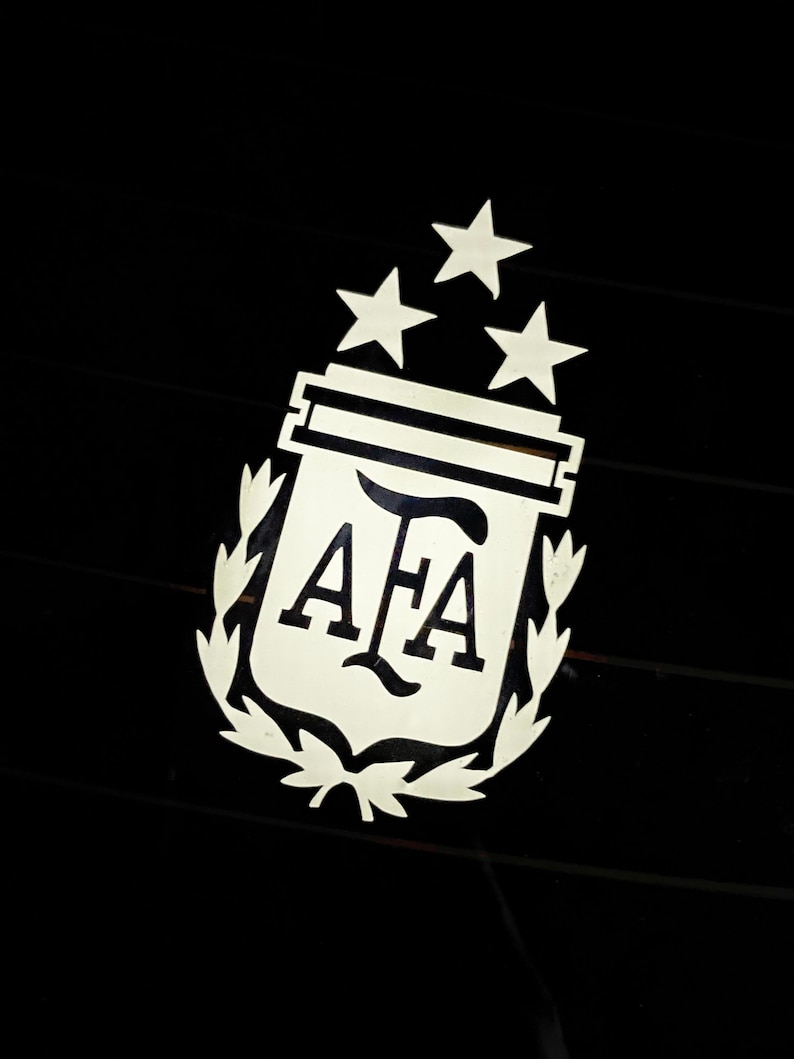 Pu&ograve; includere: Un emblema argentato con le lettere "AFA" all'interno di uno scudo, sormontato da tre stelle e circondato da una corona d'alloro. L'emblema &egrave; posto su uno sfondo nero.