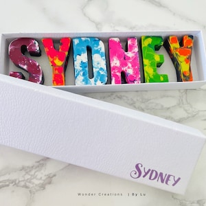 May include: A white box with the name "Sydney" spelled out in colorful, melted crayon letters. The box is open and the letters are visible inside. The box has the text "Wonder Creations | By Lu" on the bottom.