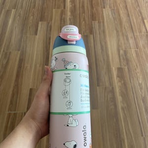 May include: A pink and blue Owala FreeSip water bottle with Snoopy graphics. The bottle has a push-button lid and a built-in straw. The bottle is 32 oz (946 ml).