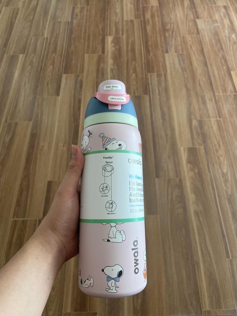 May include: A pink and blue Owala FreeSip water bottle with Snoopy graphics. The bottle has a push-button lid and a built-in straw. The bottle is 32 oz (946 ml).