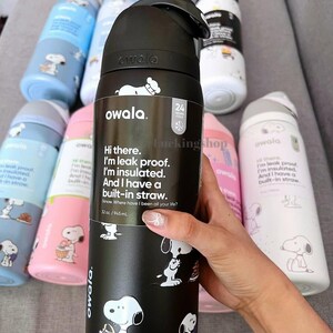 May include: A black Owala water bottle with a 32 oz capacity, featuring a Snoopy graphic and text that reads: Hi there. I'm leak proof. I'm insulated. And I have a built-in straw. Several other colorful water bottles with Snoopy designs are visible.