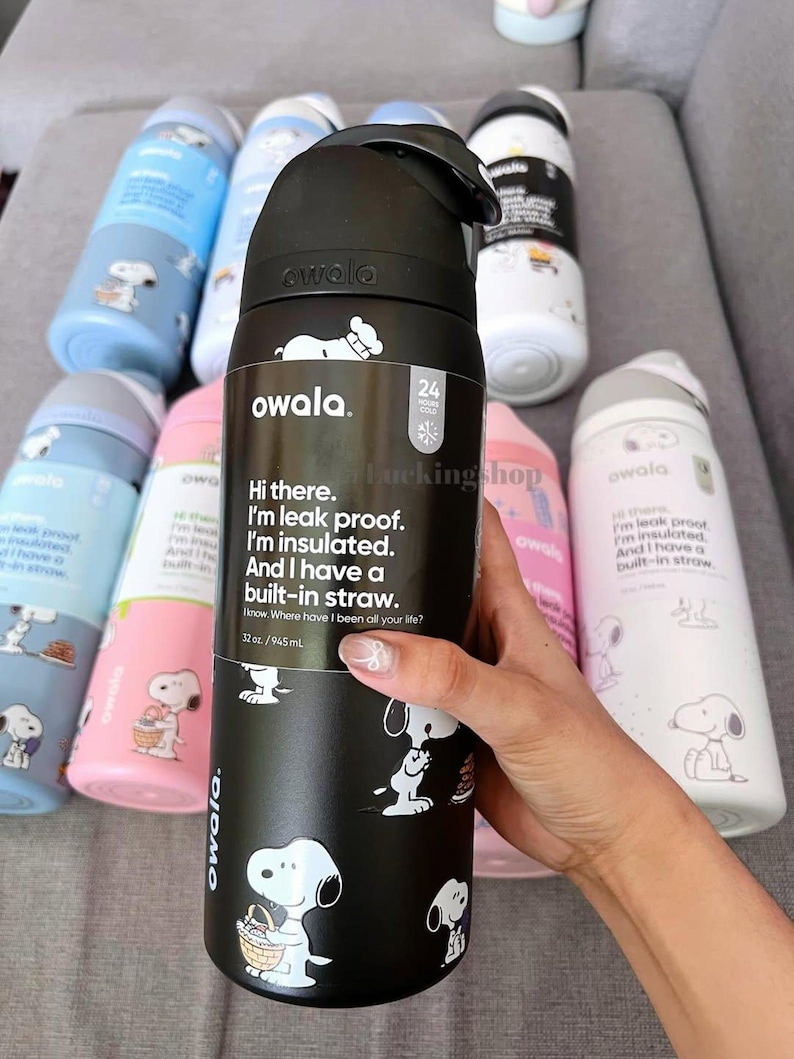 May include: A black Owala water bottle with a 32 oz capacity, featuring a Snoopy graphic and text that reads: Hi there. I'm leak proof. I'm insulated. And I have a built-in straw. Several other colorful water bottles with Snoopy designs are visible.