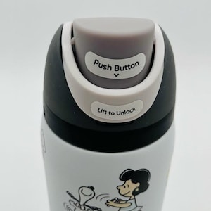 Puede incluir: Botella de agua aislada blanca y negra con un dise&ntilde;o de dibujos animados de Snoopy y Lucy. La tapa tiene un bot&oacute;n gris y el texto "Push Button" y "Lift to Unlock".