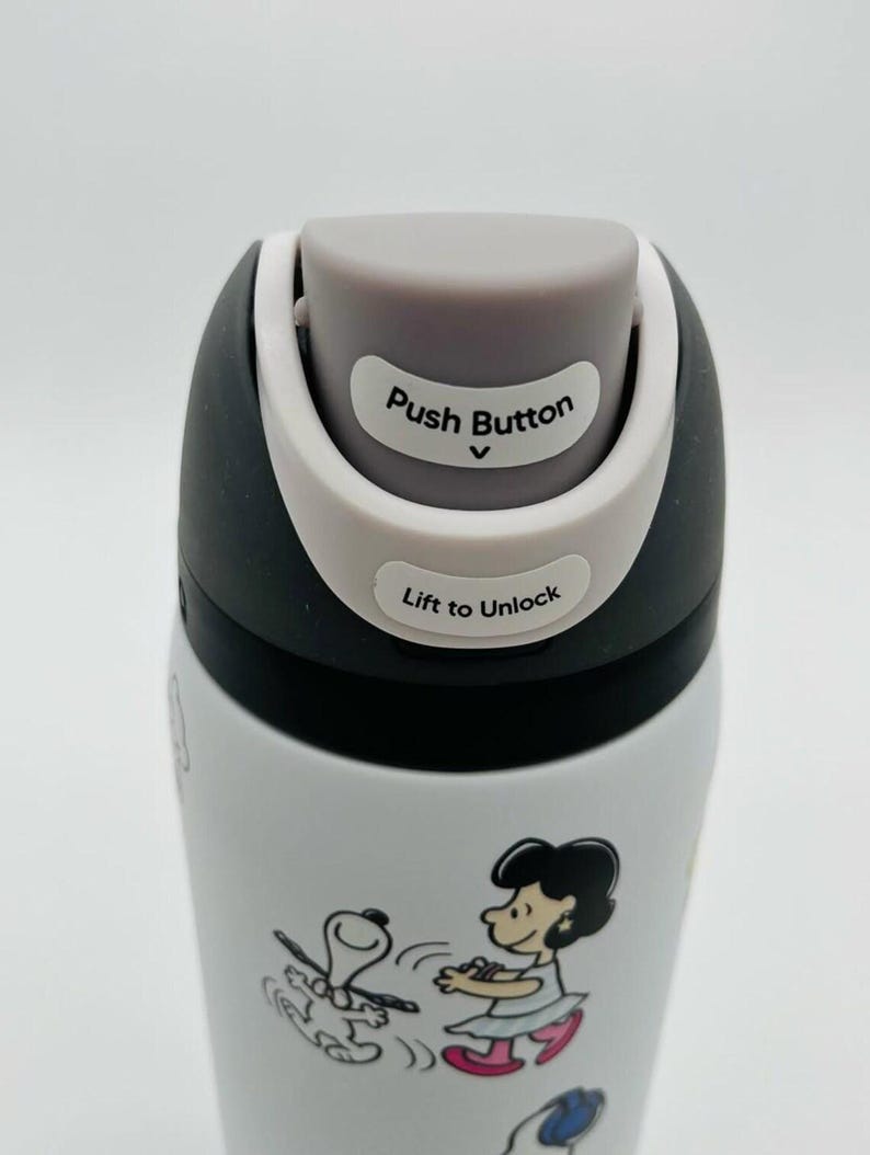Puede incluir: Botella de agua aislada blanca y negra con un dise&ntilde;o de dibujos animados de Snoopy y Lucy. La tapa tiene un bot&oacute;n gris y el texto "Push Button" y "Lift to Unlock".