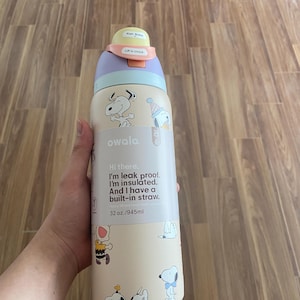 May include: A beige Owala water bottle featuring Snoopy illustrations. The bottle has a purple, light blue, and yellow lid. Text on the bottle reads "Hi there. I'm leak proof. I'm insulated. And I have a built-in straw." It holds 945ml.