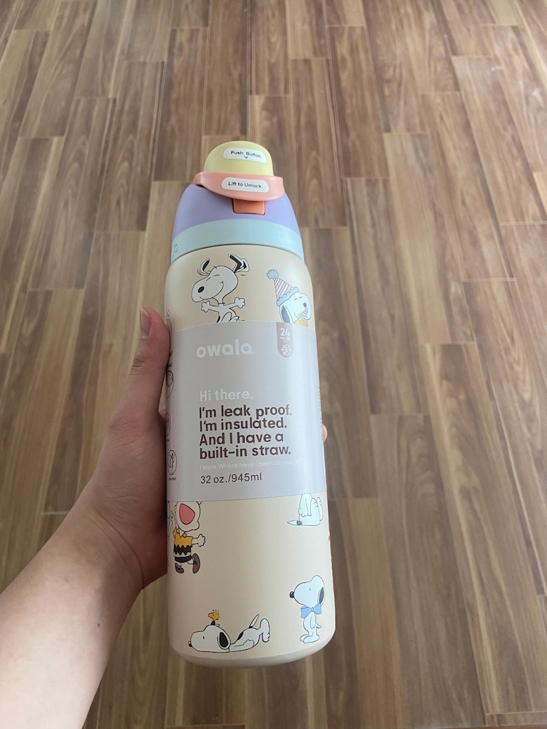 May include: A beige Owala water bottle featuring Snoopy illustrations. The bottle has a purple, light blue, and yellow lid. Text on the bottle reads "Hi there. I'm leak proof. I'm insulated. And I have a built-in straw." It holds 945ml.
