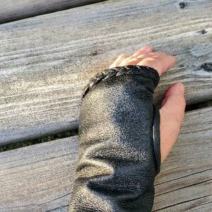 May include: Black leather wrist cuff with a braided detail and a black leather strap. The cuff is worn on a person's left wrist.