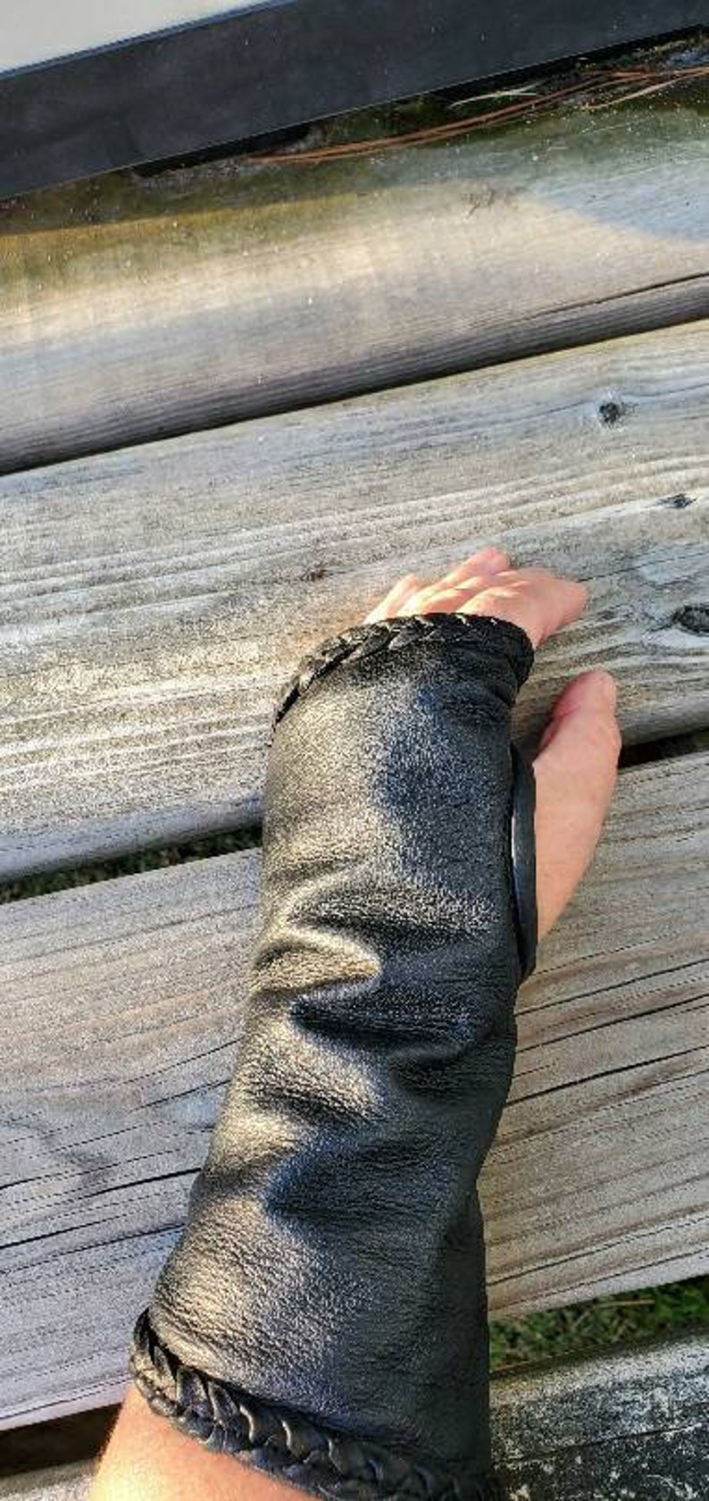 May include: Black leather wrist cuff with a braided detail and a black leather strap. The cuff is worn on a person's left wrist.