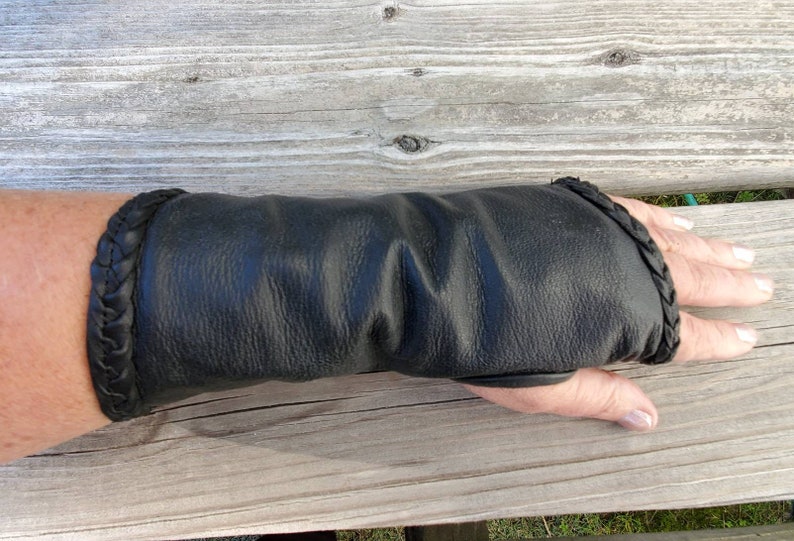 May include: Black leather wrist warmer with a braided detail. The warmer is worn on a person's left wrist.