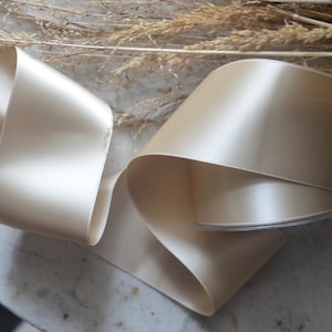 May include: A roll of ivory satin ribbon, folded and displayed on a white surface. The ribbon is smooth and shiny, with a subtle sheen.