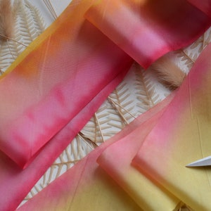 May include: A close-up of a yellow, orange, and pink ombre ribbon. The ribbon is folded and arranged on a white surface with dried fern leaves.
