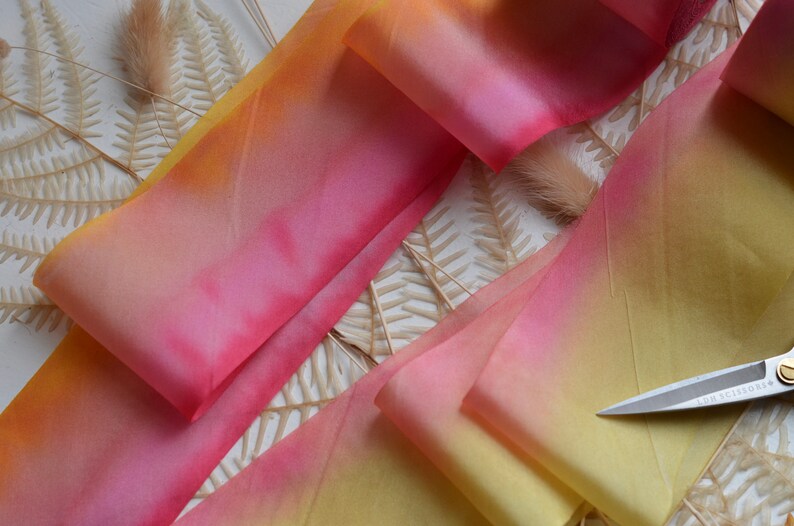 May include: A close-up of a yellow, orange, and pink ombre ribbon. The ribbon is folded and arranged on a white surface with dried fern leaves.