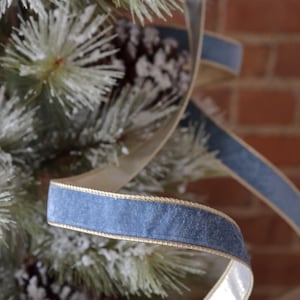 Op de afbeelding: Close-up van een blauw fluwelen lint met gouden rand, gewikkeld rond een bevroren dennentak. De textuur en kleur van het lint contrasteren met het groen en wit van de dennennaalden en een bruine dennenappel. De achtergrond is een rode bakstenen muur.