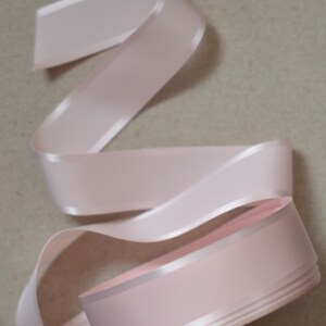 May include: A pale pink satin ribbon with a slightly textured surface. The ribbon is folded and coiled, showing the edges and the smooth satin finish.