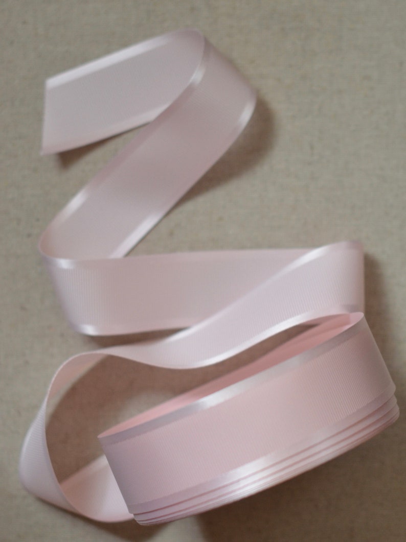 May include: A pale pink satin ribbon with a slightly textured surface. The ribbon is folded and coiled, showing the edges and the smooth satin finish.