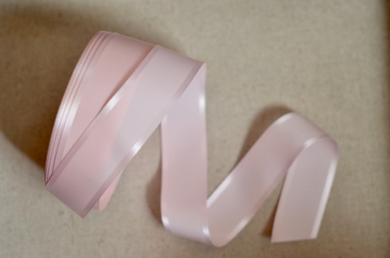 May include: A pale pink satin ribbon with a slight sheen. The ribbon is folded and twisted into a decorative shape.