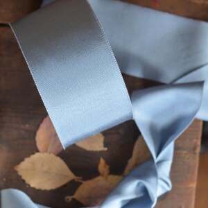 May include: A light blue satin ribbon with a textured surface. The ribbon is folded and arranged on a wooden surface with dried leaves.