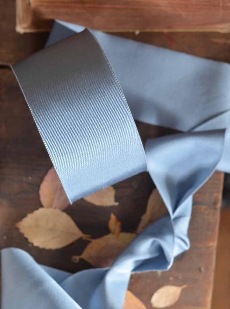 May include: A light blue satin ribbon with a textured surface. The ribbon is folded and arranged on a wooden surface with dried leaves.